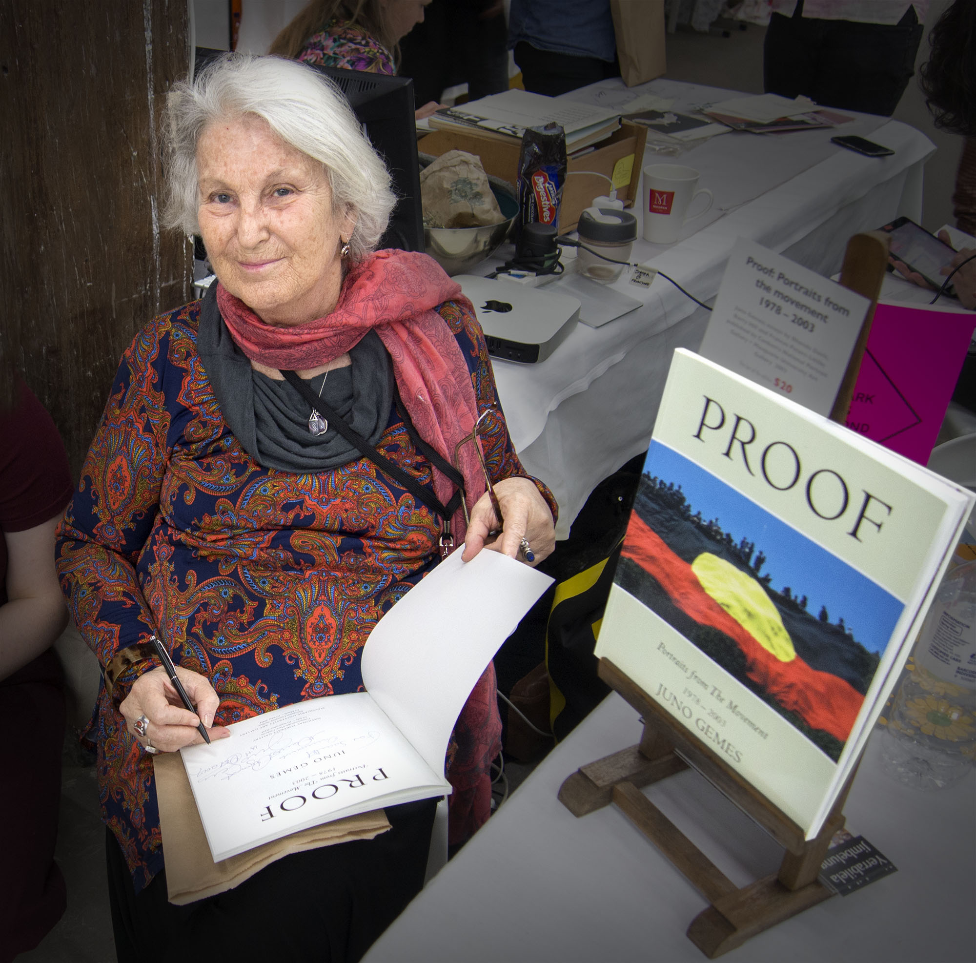 An elderly woman with gray hair, wearing a patterned blouse and a red scarf, is sitting at a table. She is signing a book titled 'PROOF' while holding a pen and a blank page. A display of the book is visible on the table next to her.